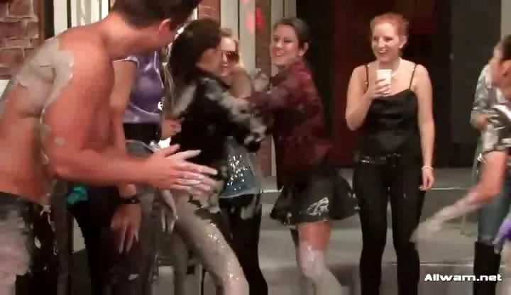 A group of girls are standing around a pool which is filled with mud. Two of them get into the pool and start wrestling each other, throwing each other over. After a few minutes both of them are covered in grey mud.